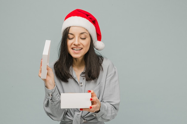 Foto de uma fofa mulher sorridente com chapéu de Natal está abrindo o presente. Veste camisa cinza, fundo de cor cinza isolado.