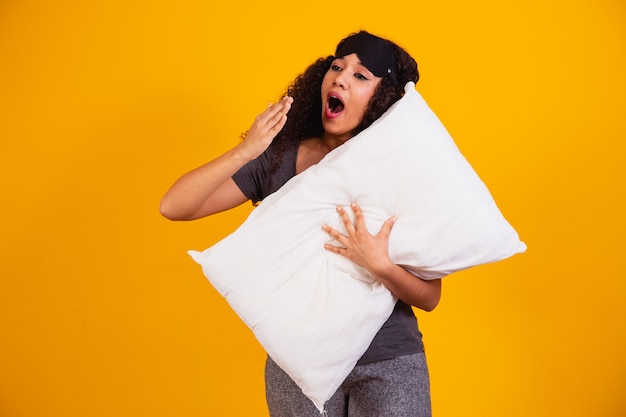 Foto de uma garota afro, abraçando o travesseiro no fundo amarelo, bocejando de sono.