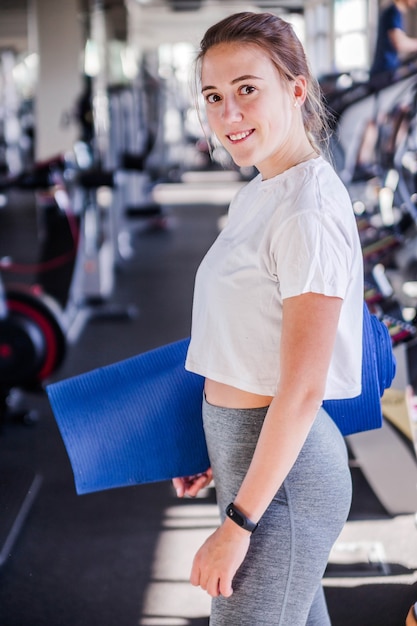 Foto de uma garota com um tapete de ginástica nas mãos dela posa no fundo do ginásio. Sorrindo e olhando para a câmera