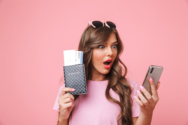 Foto de uma garota de 20 anos de viagem surpresa olhando para o celular em choque, segurando o passaporte e as passagens aéreas nas mãos, isolada sobre um fundo rosa
