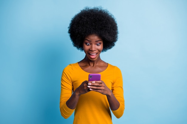 Foto de uma garota de pele negra espantada e surpresa, mantendo o sorriso de mensagens de texto no smartphone isolado em um fundo de cor azul brilhante