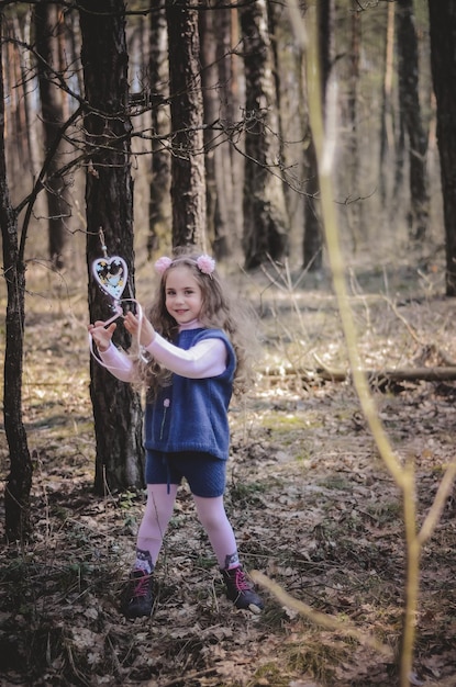 Foto de uma garota sorridente em uma bandana brincando com um coração de madeira na floresta