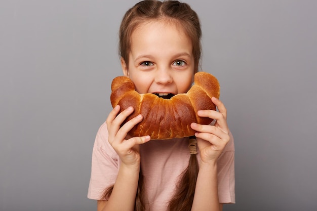 Foto de uma garotinha alegre com cabelo escuro mordendo uma grande e saborosa massa de bagel isolada sobre um fundo cinza com um lanche saboroso comendo uma deliciosa padaria caseira