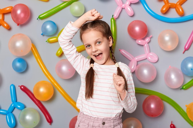 Foto de uma garotinha animada e alegre com tranças vestindo roupas casuais, punhos cerrados, braços levantados, comemorando o aniversário dançando em pé contra a parede cinza com balões coloridos