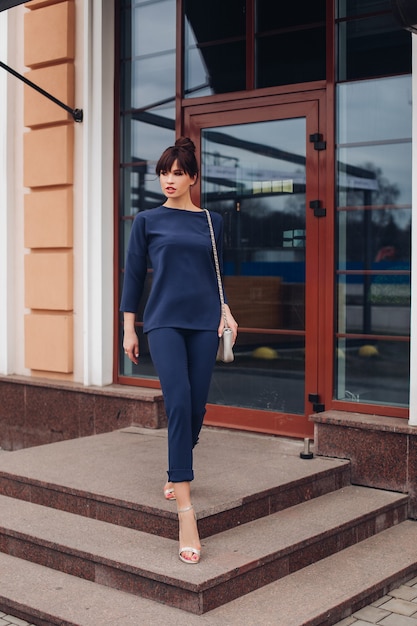 Foto de uma jovem adorável mulher branca com cabelo escuro, blusa azul escuro e calça azul escuro, sapatos brancos com bolsa branca