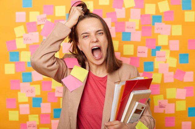 Foto de uma jovem adulta louca e cansada segurando papéis posando contra a parede amarela com adesivos coloridos gritando muito alto por estar exausta sente cansaço do trabalho ou estudando rasga o cabelo na cabeça