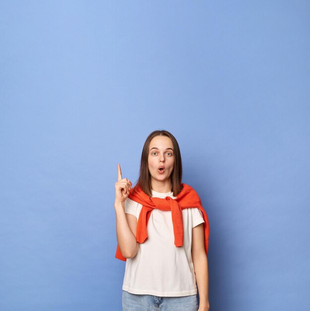 Foto de uma jovem adulta maravilhada, vestida de camiseta branca e suéter laranja amarrado sobre os ombros, em pé contra a parede azul, apontando para cima no espaço da cópia para propaganda