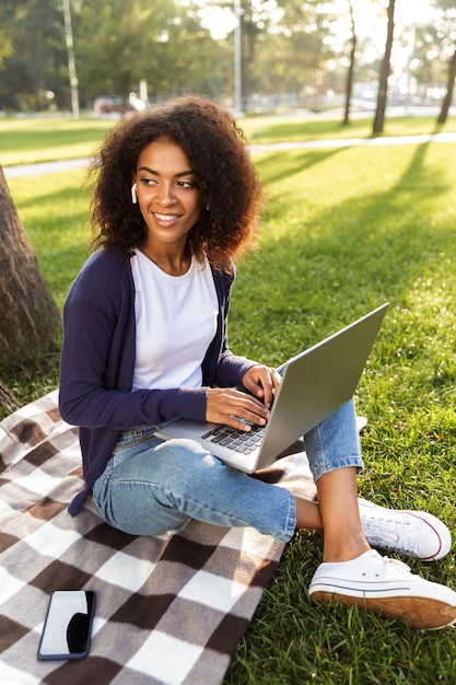 Foto de uma jovem africana sentada ao ar livre no parque, usando o computador portátil, ouvindo música com fones de ouvido.