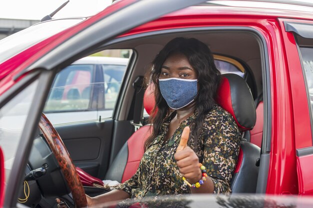 Foto de uma jovem afro-americana com uma máscara facial protetora mostrando os polegares para cima enquanto dirige