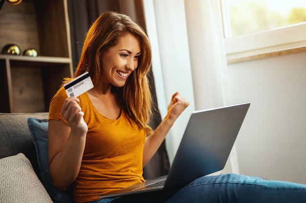 Foto de uma jovem alegre fazendo compras on-line em seu laptop enquanto está sentado no sofá em casa.