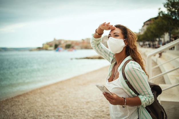 Foto de uma jovem atraente e feliz usando máscara protetora N95 e usando smartphone enquanto desfruta de férias na praia durante o COVID-19.