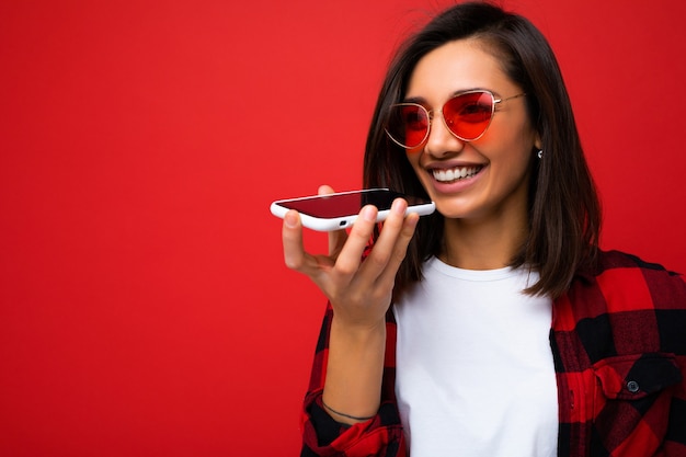 Foto de uma jovem atraente, positiva e bonita vestindo roupa casual elegante, posing isolado no fundo com um espaço vazio segurando na mão e usando a gravação de voz sms de telefone celular