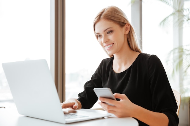 Foto de uma jovem atraente trabalhadora sentada no escritório enquanto usa o computador laptop e bate-papo por telefone. olha para o computador