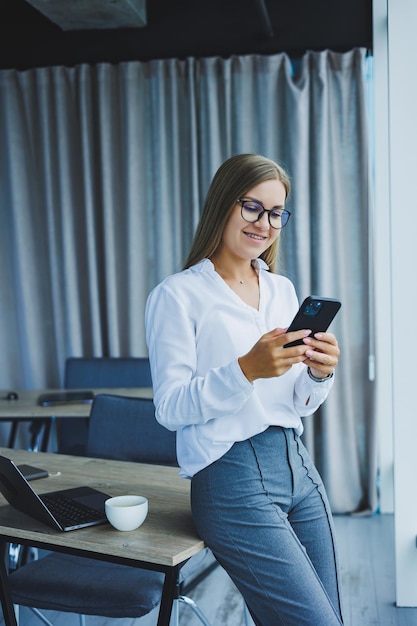 Foto de uma jovem bem sucedida em uma camisa sorrindo e trabalhando em um laptop enquanto fala ao telefone em um escritório moderno com grandes janelas Trabalho remoto com café