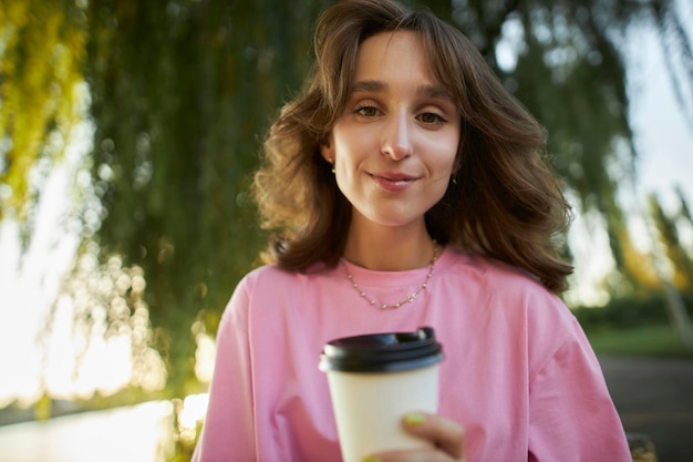 Foto de uma jovem bonita e charmosa em camiseta rosa, no parque, com um sorriso segurando uma xícara de café de papel.