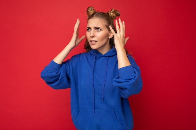 Foto de uma jovem bonita loira atônita com dois chifres e emoções sinceras, vestindo um capuz azul brilhante moderno isolado sobre um fundo vermelho com espaço de cópia