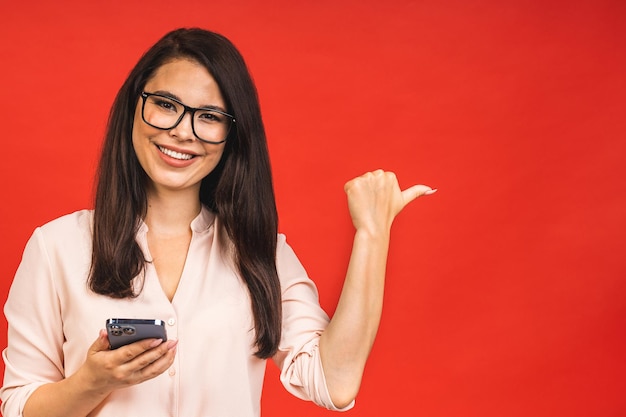 Foto de uma jovem de negócios bonita e alegre usando telefone celular isolado sobre fundo vermelho espaço para texto apontando o dedo