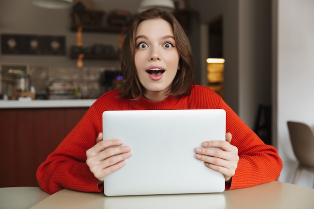 Foto de uma jovem encantada de 20 anos usando uma blusa surpresa e segurando um laptop prateado nas mãos, enquanto está sentada em um café