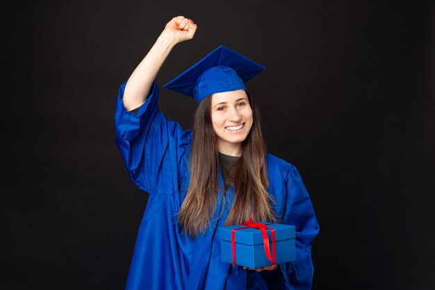 Foto de uma jovem estudante com um manto azul comemorando o sucesso e segurando uma caixa de presente