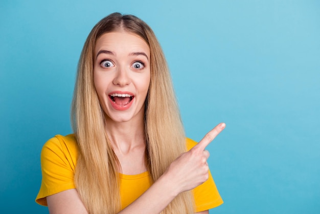 Foto de uma jovem excitada vestida de camisa sorrindo apontando o espaço vazio do dedo isolado de fundo azul