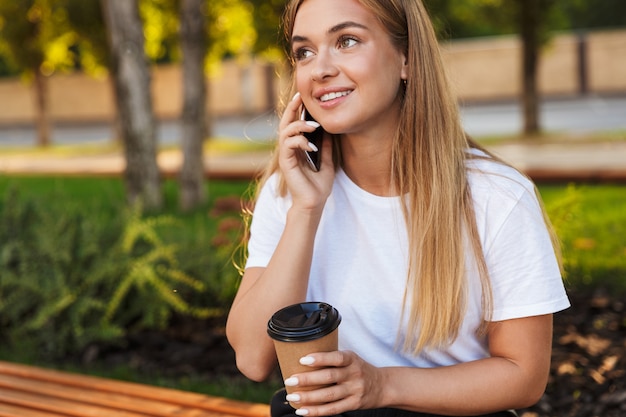 Foto de uma jovem feliz alegre positiva sentar no parque natural falando por telefone celular, bebendo café.