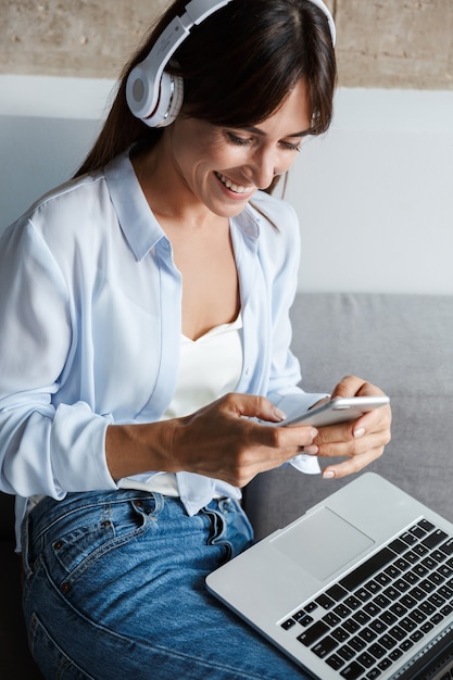 Foto de uma jovem feliz positiva alegre dentro de casa em casa usando música laptop com fones de ouvido, sente-se no sofá na sala de estar conversando por telefone celular.