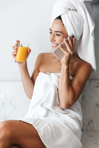 Foto de uma jovem incrível mulher feliz na toalha após o procedimento de spa tomar banho dentro de casa em casa na cozinha, bebendo suco, falando por telefone celular.