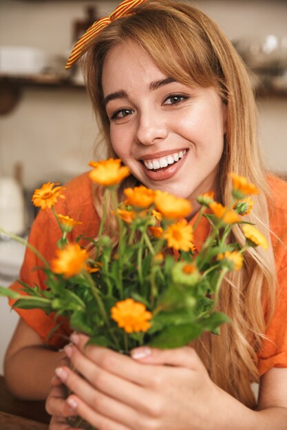 Foto de uma jovem loira sorridente alegre na cozinha segurando lindas flores nas mãos.