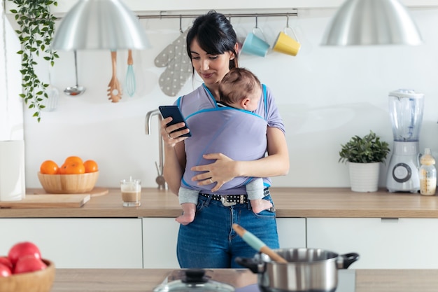 Foto de uma jovem mãe com o bebê na tipóia usando seu telefone celular na cozinha em casa.