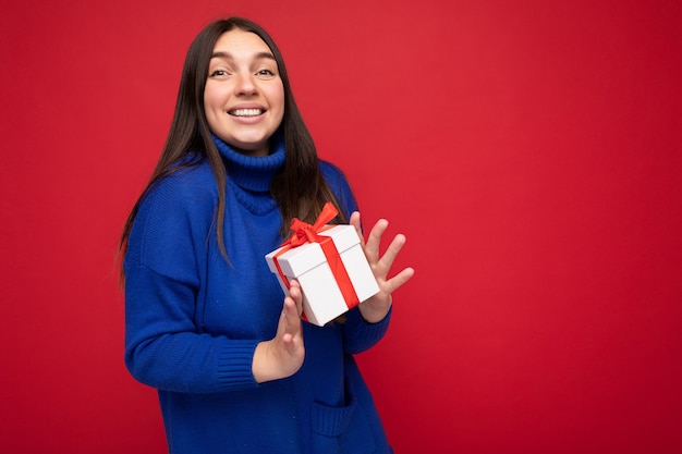 Foto de uma jovem morena atraente e feliz, isolada sobre a parede de fundo vermelho, usando uma blusa casual azul, segurando uma caixa de presente branca e olhando para a câmera