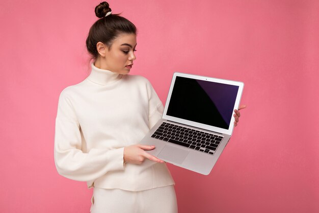 Foto de uma jovem morena bonita com cabelo escuro recolhido, suéter branco, segurando o laptop do computador e olhando para o netbook isolado no fundo da parede rosa.