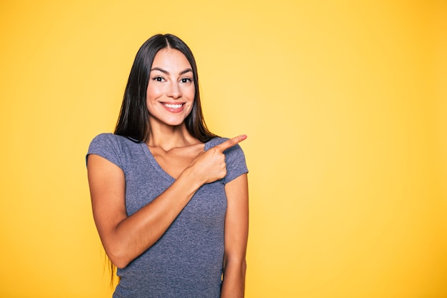 Foto de uma jovem morena feliz e animada apontando para longe e sorrindo isolada no banner de fundo amarelo