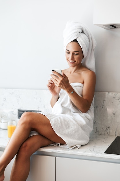 Foto de uma jovem mulher alegre e feliz na toalha após o procedimento de spa tomar banho dentro de casa em casa na cozinha usando telefone celular.