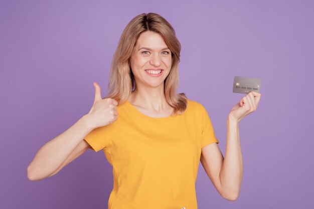 Foto de uma jovem mulher atraente feliz sorriso positivo segurar cartão de crédito mostrar polegar para cima como um anúncio legal isolado em um fundo de cor violeta