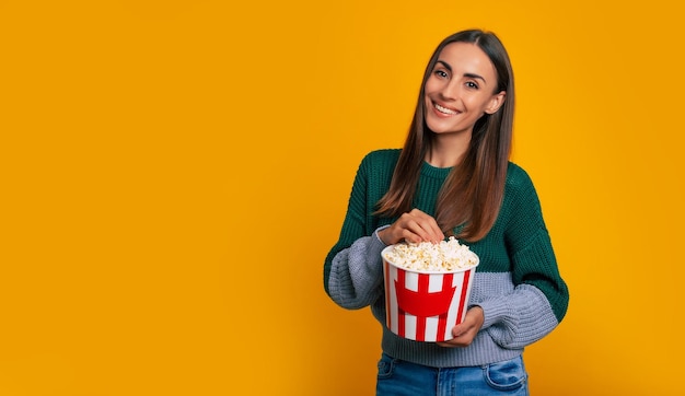 Foto de uma jovem mulher bonita e feliz com um suéter enquanto segura um grande balde de pipoca para assistir o filme no cinema e olhar para a câmera com um sorriso