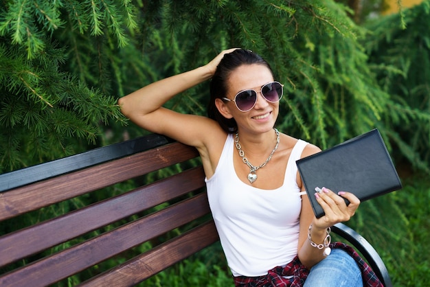 Foto de uma jovem mulher bonita sentada num banco de madeira no parque com o bloco de notas preto na mão em cas ...