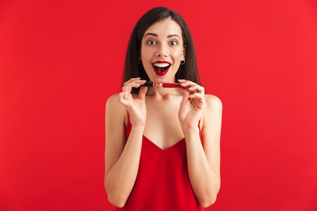 Foto de uma jovem mulher feliz e animada posando isolada segurando brilho labial, fazendo maquiagem.