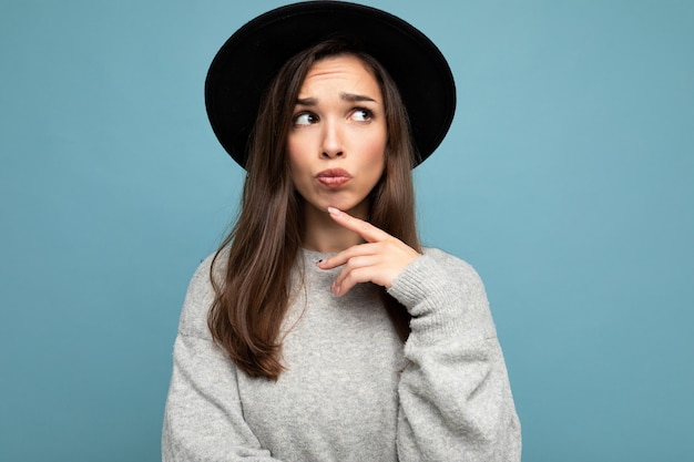 Foto de uma jovem mulher morena bonita atraente pensativa com emoções sinceras, vestindo um suéter cinza casual e um chapéu preto isolado sobre um fundo azul com espaço livre e pensamento