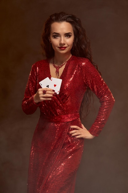 Foto de uma jovem mulher morena bonita caucasiana em um vestido de noite vermelho brilhante tem cartas em uma mão, mostra um par de ases, segurando uma mão no quadril. fundo marrom, cassino, indústria de jogos de azar