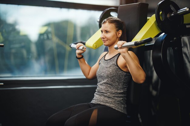 Foto de uma jovem musculosa em roupas esportivas malhando na máquina de ginástica. Ela está bombeando o músculo do ombro com peso pesado.