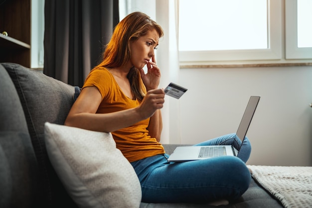 Foto de uma jovem preocupada fazendo compras on-line em seu laptop enquanto está sentado no sofá em casa.