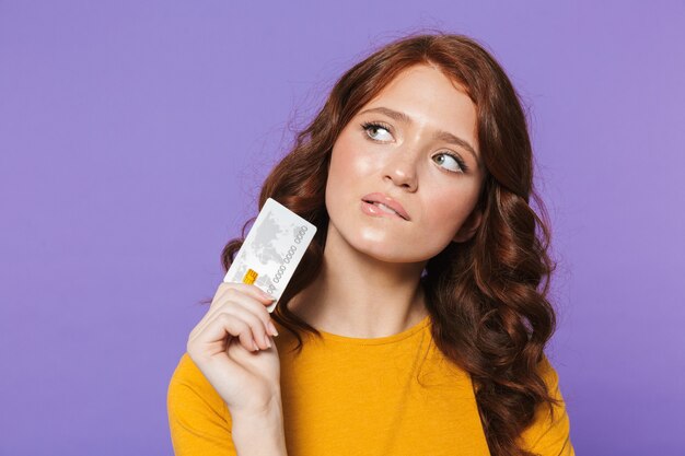 Foto de uma jovem ruiva vestindo roupas amarelas segurando um cartão de crédito de plástico e olhando para cima sobre o roxo