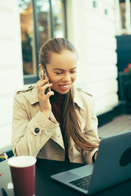 Foto de uma jovem sentada em um café falando ao telefone na frente de um computador com um café