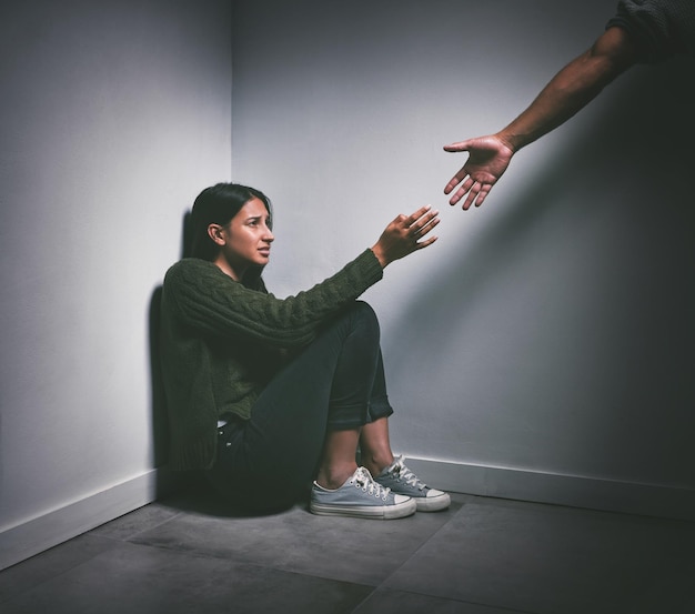 Foto de uma jovem sentada no canto de uma sala escura com uma mão estendida para ajudá-la