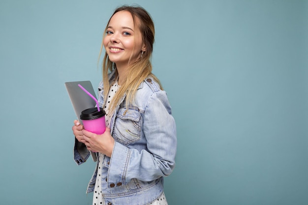 Foto de uma jovem sorridente loira vestindo uma jaqueta jeans isolada sobre um fundo azul carregando