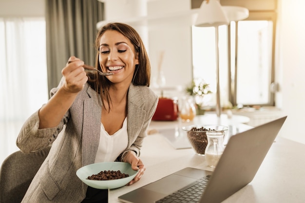 Foto de uma jovem sorridente tomando café da manhã enquanto usa o laptop em casa.