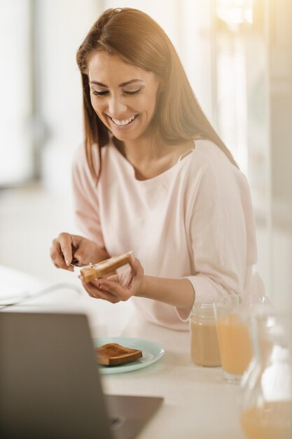 Foto de uma jovem sorridente usando laptop enquanto preparava o café da manhã para si mesma em sua cozinha.