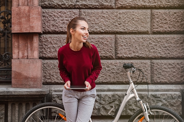 Foto de uma jovem sorridente, vestida com um suéter, em pé perto de sua bicicleta na rua e usando um tablet