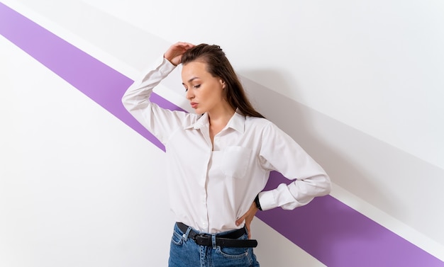 Foto de uma jovem vestida com uma camisa branca. De pé ainda isolado sobre uma parede branca com faixa violeta.