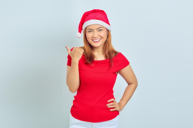 Foto de uma linda garota asiática com chapéu de Natal com expressão facial sorridente e apontando para o lado